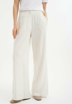 Vrouw die witte brede pijpen draagt met dunne verticale zwarte strepen, een wit overhemd en beige schoenen, staand met één hand in de zak.