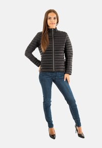 Giacca a vento nera con design a imbottitura orizzontale, colletto alto, zip grigia, abbinata a jeans blu scuro e tacchi neri.