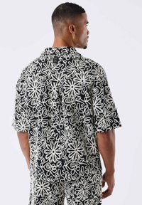 Camisa de manga corta en negro con patrones florales y abstractos en blanco, cuenta con un cuello plano y una etiqueta en la parte posterior del cuello.