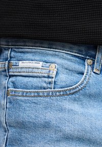 Κοντινή λήψη τσεπής ανοιχτού μπλε τζιν με ετικέτα Marc O'Polo Denim και μεταλλικά καρφιά, σε συνδυασμό με μαύρο υφασμάτινο ένδυμα.