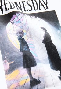 Camiseta gráfica con dos personajes vestidos de negro, uno con chaqueta a rayas y falda. El fondo presenta un diseño colorido de vidrieras.