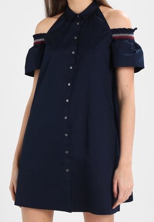 Femme portant une robe bleu marine à boutons sur le devant, épaules dénudées, manches courtes avec une bordure brodée sur le haut des bras.