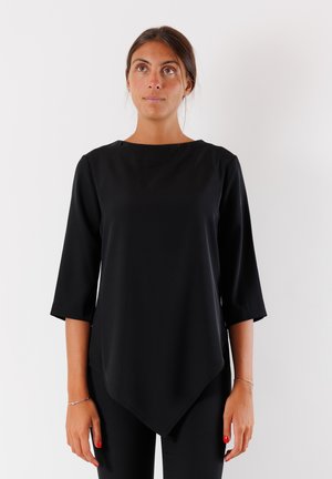 Top nero a maniche lunghe con leggero scollo a barca e orlo asimmetrico curvo. Tessuto morbido e leggero. Indossato con pantaloni neri.