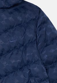 Tamno plavi dukserica s kviltanim uzorkom koja sadrži ponavljajući reljefni logo "Nike" i teksturiranu površinu. Uključuje kapuljaču i glatku dodirnu površinu.