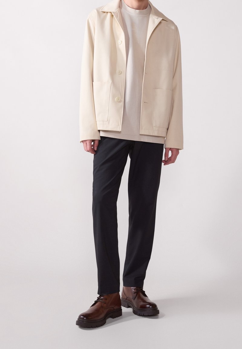 Homme portant une veste beige et une chemise, un pantalon noir et des chaussures en cuir marron, debout sur un fond blanc.