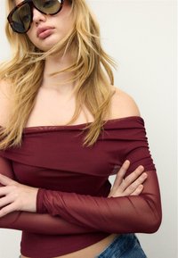 Weinrotes schulterfreies Oberteil mit langen durchsichtigen Ärmeln, kombiniert mit großen Sonnenbrillen im Schildpatt-Design, getragen vor neutralem Hintergrund.