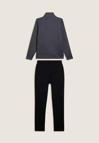 Giacca grigia scura a maniche lunghe con colletto abbinata a pantaloni neri a gamba dritta, distesi su uno sfondo beige chiaro.