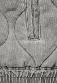 Primer plano de una tela gris acolchada con formas ovaladas cosidas, una correa vertical con bucles y elastico recogido en el borde inferior.