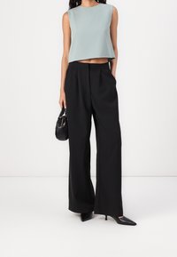 Donna con top corto verde salvia senza maniche, pantaloni neri a gamba larga a vita alta, tacchi neri a punta e una borsa nera in mano.