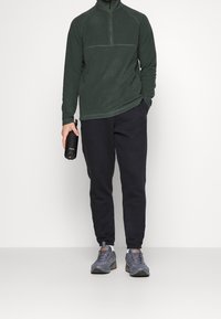 Osoba v tmavozeleném fleece s zipem a černých teplácích, která drží černou láhev S'well, má na sobě šedé tenisky s bílými ponožkami.