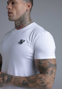 Hvid kortærmet t-shirt med tætsiddende design, der har en lille sort logo på venstre brystområde og en glat, blød tekstur.