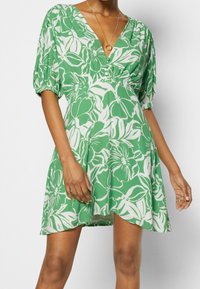 Zelené šaty s bílým květinovým vzorem, hlubokým V-výstřihem, krátkými nabíranými rukávy a elastickým pasem. Lehká tkanina s volným střihem.