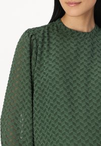 Haut vert texturé avec un motif géométrique en chevrons, un col côtelé et des manches bouffantes en tissu léger.