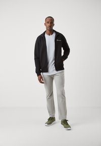 Chaqueta negra con cremallera, camiseta gris, pantalones de chándal grises y zapatillas verdes. Presenta logos en la chaqueta y los pantalones. Tela suave, diseño casual.