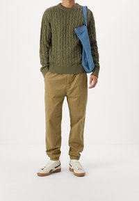 Olivově zelený svetr s pleteným vzorem, khaki tapered kalhoty s elastickými manžetami a bílé tenisky s černými detaily. Modrá látková kabelka přes rameno.