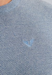 Hellblauer Strickpullover mit einem strukturierten Muster und einem kleinen, blauen gestickten Emblem auf der Brust. Rundhalsausschnitt.