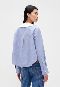 Koszula w paski z długim rękawem w niebiesko-białym kolorze, wykonana z bawełny, z okrągłym kołnierzykiem, plisą z tyłu i rozcięciami po bokach. Mankiety zapinane na guziki i luźny krój.