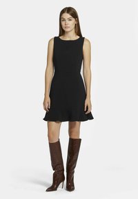 Schwarzes ärmelloses Kleid mit ausgestelltem Saum, aus glattem Stoff gefertigt. Kombiniert mit kniehohen, braunen Stiefeln aus Krokodillederoptik mit spitzer Zehe.