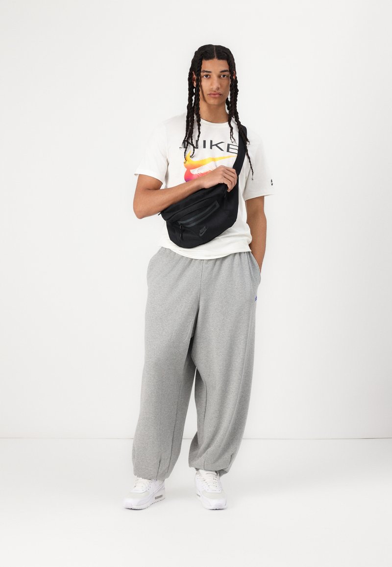 Czarna torba na ramię Nike, szare dresy i biały t-shirt z kolorowym logo Nike. Casualowy strój w luźnym fasonie i sneakersy.
