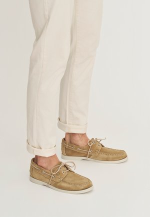 Benedenbenen met opgerolde beige broekspijpen en lichtbruine suède bootschoenen met veters, weergegeven in een staande positie tegen een neutrale achtergrond.