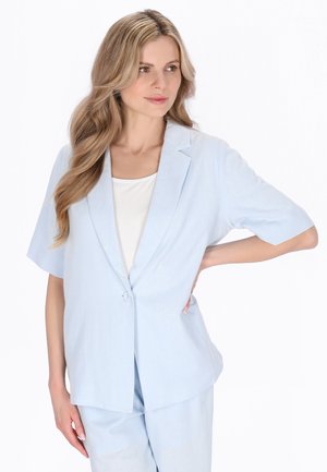 Femeie cu păr blond lung, purtând un sacou bleu cu mâneci scurte și pantaloni asortați, stând cu o mână pe șold.