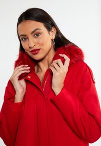 Rode parka-jack met een bontgevoerd capuchon, gouden rits en geribde manchetten. De stof heeft een gladde textuur en beschikt over een ontspannen pasvorm.