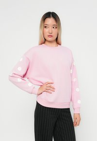 Rosa sweatshirt med långa ärmar, har vita blommiga applikationer på ärmarna, rundad halsringning och en avslappnad passform. Kombination med svarta randiga byxor.