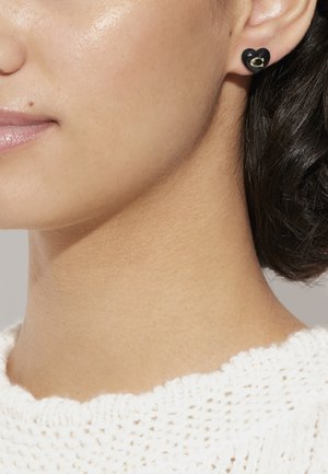 Orecchini a forma di cuore neri con un accento dorato, indossati all'orecchio di un modello. La texture morbida e la finitura lucida sono visibili su una pelle chiara.
