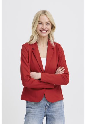 Femme blonde souriante avec assurance, les bras croisés, portant un blazer rouge sur un haut blanc et un jean bleu clair, sur un fond uni.