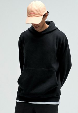 Schwarzer Kapuzenpullover mit Kängurutasche, aus weichem Stoff. Kombiniert mit einer hellrosa Baseballmütze mit einem Logo.