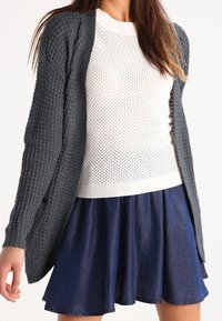Stickad mörkgrå cardigan med öppen framsida, strukturerat mönster, tillsammans med en vit perforerad tröja och en marinblå utsvängd jeanskjol.