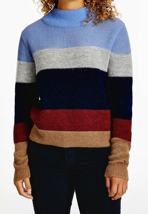 Personne portant un pull côtelé avec des rayures horizontales bleu, gris, marine, rouge et beige, associé à un pantalon sombre.