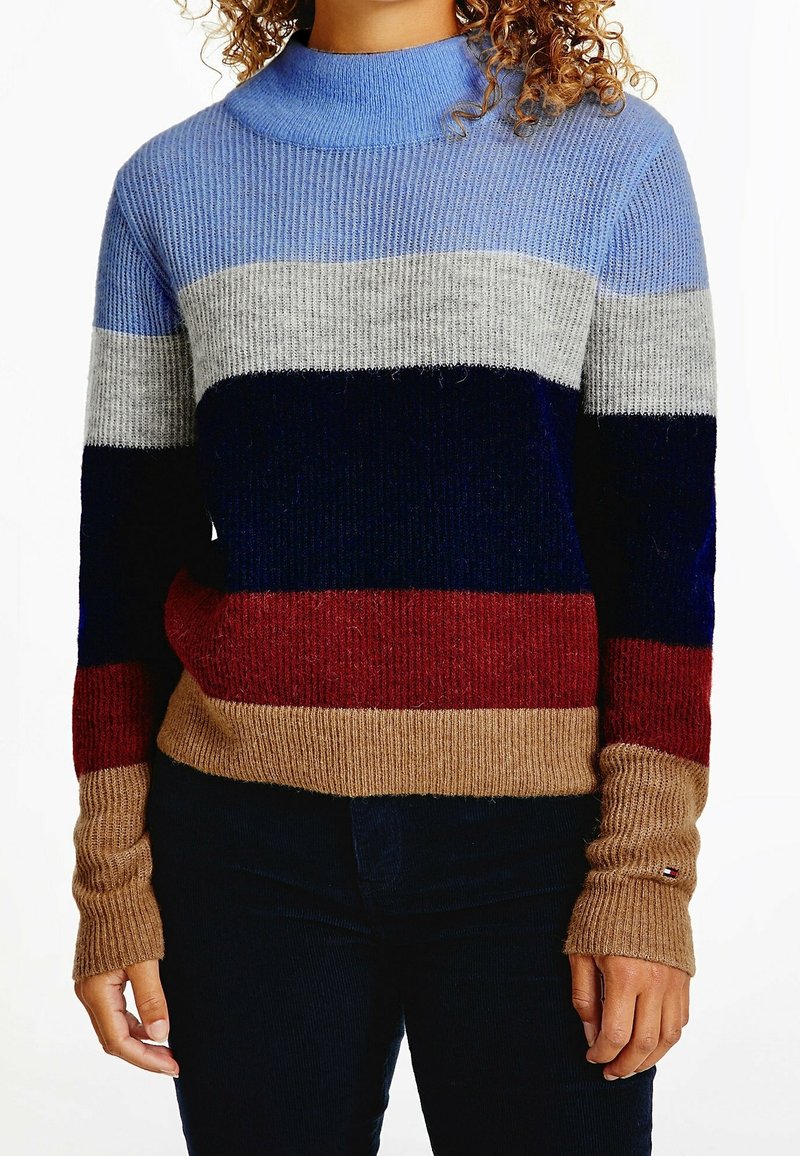 Personne portant un pull côtelé avec des rayures horizontales bleu, gris, marine, rouge et beige, associé à un pantalon sombre.