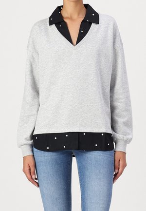 Kobieta w jasnoszarym swetrze z dekoltem w serek, nałożonym na czarną koszulę w białe kropki, w jasnoniebieskich dżinsach, stojąca na jednolitym tle.