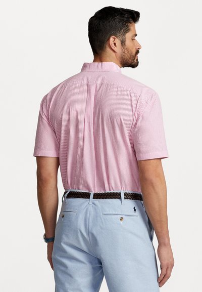 Īss piedurkņu krekls ar rozā un baltu svītru rakstu, auduma tekstūra ir redzama; saskaņots ar gaiši ziliem biksēm un brūnu austu jostu.