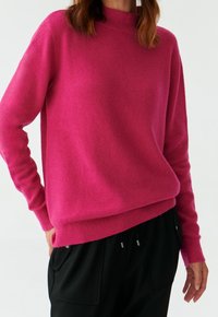 Fuchsia Wolle-Mix Pullover mit hohem Kragen, geripptem Saum und Bündchen; lockerer Schnitt; kombiniert mit schwarzen Hosen, die eine glatte Textur zeigen.