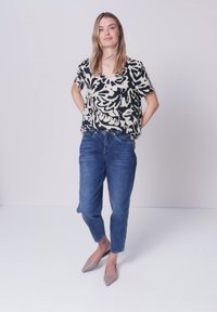 Jeune femme debout, les mains dans le dos, portant un chemisier à manches courtes noir et blanc à motifs, un jean bleu et des ballerines pointues beiges sur un sol blanc.