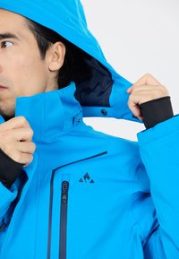 Blå vattentät jacka med hög krage och justerbar huva. Har svarta dragkedjor och detaljer, mjukt innertyg och en ficka med dragkedja på bröstet.