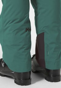 Groene waterdichte skibroek met HELLY TECH®-merk, met een zwart paneel op het onderbeen, gecombineerd met zwarte skibotten.