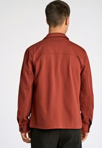 Rostfarbener Mantel aus einem weichen Stoff mit Kragen, knöpfbaren Manschetten und geradem Schnitt. Minimalistisches Design, keine Muster oder Verzierungen.