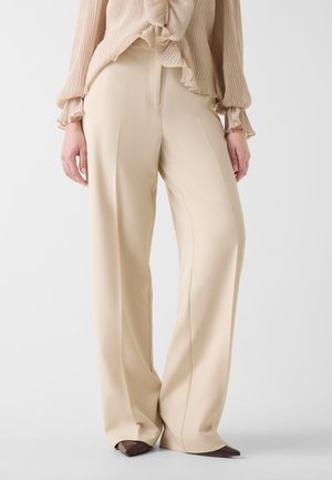Donna che indossa pantaloni beige a gamba larga con piega sul davanti, abbinati a una camicetta beige chiaro a maniche lunghe e scarpe marroni con punta.
