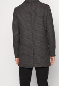 Cappotto grigio a scacchi con un design sartoriale, maniche lunghe, colletto a punta e taglio dritto. La texture del tessuto è liscia e strutturata.