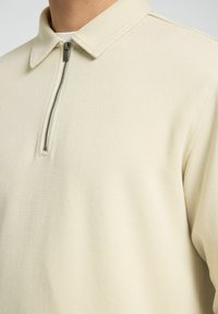 Polo cremoso con cremallera, fabricado en un tejido texturizado, que presenta un cuello plano y un detalle de cremallera plateada en el cuello.