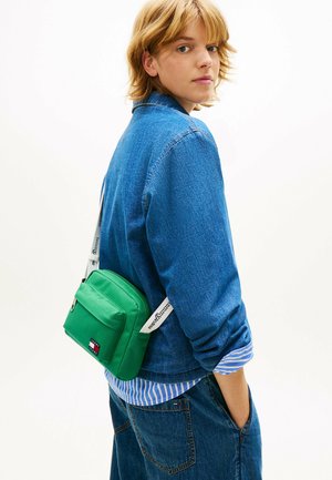 Persona joven con una chaqueta y pantalones de mezclilla azul, llevando una bolsa cruzada verde con una correa blanca sobre un fondo blanco liso.