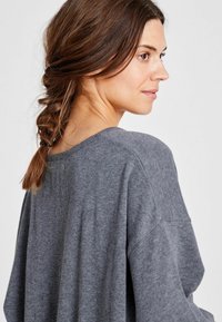 Sivi pleteni pulover z ohlapnim krojem, širokim okroglim izrezom in spuščenimi rameni. Model ima spleteno frizuro, ki prikazuje zadnji del dizajna.
