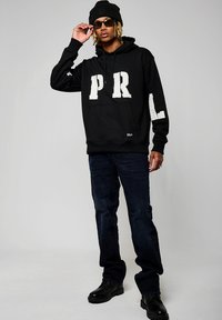 Haut à capuche noir avec les lettres blanches en bloc "P" et "R" sur le devant et "L" sur une manche. Un jean sombre et des chaussures en cuir noires complètent le look.