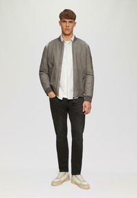 Grijze bomberjack met ribgebreide mouwen en kraag, wit gestreept shirt, zwarte jeans en beige sneakers, staande voor een effen achtergrond.