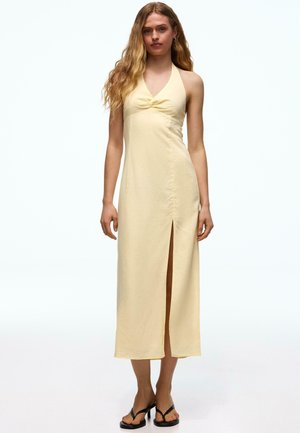 Frau mit langen, welligen Haaren, die ein ärmelloses, hellgelbes Neckholder-Kleid mit einem Schlitz vorne trägt und schwarze Sandalen mit Absatz anhat.