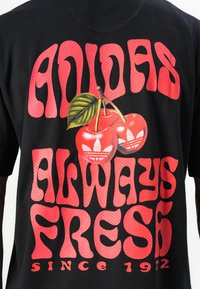 T-shirt noir graphique présentant un texte rouge audacieux "ADIDAS TOUJOURS FRAIS DEPUIS 1972" et un graphique de cerise coloré avec une feuille verte.