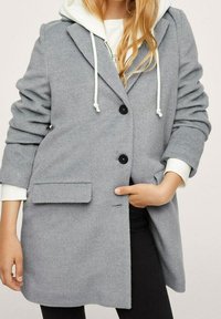 Šedý kabát z vlněné směsi s přizpůsobeným designem, jedním knoflíkem vpředu, uzavřením na dva knoflíky a náprsní kapsou. Obsahuje kapuci na vnitřní straně.